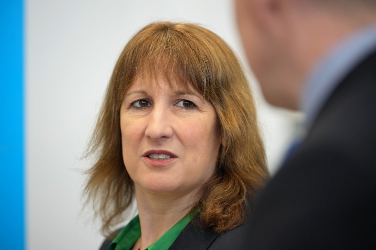 Rachel Reev addressing an audience at a business conference podium, wearing a blue blazer, discussing economic trends.