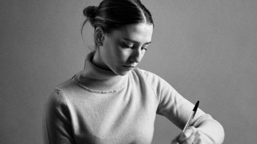 Young woman in turtleneck writing intently with a pen, dark bun hairstyle, black and white image