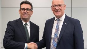 Andy Burnham and John Swinney shaking hands, both wearing dark suits and ties, in a professional setting.