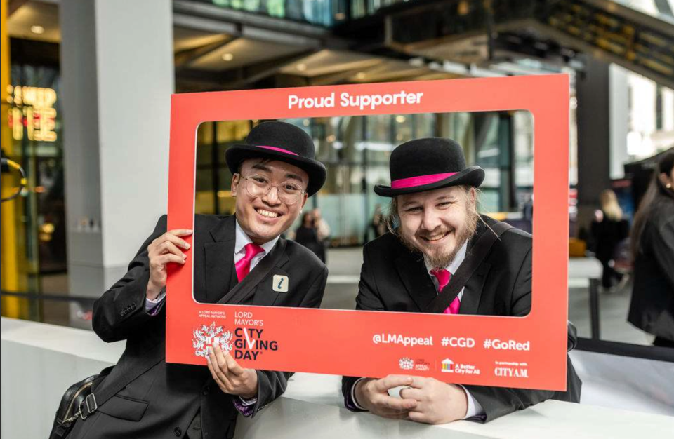 Two men in black suits and bowler hats with pink bands and ties, smiling through a red City Giving Day frame.