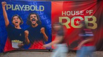 Blue and red banners featuring Playbold and House of RCB with cheering fans, blurred figures passing by.
