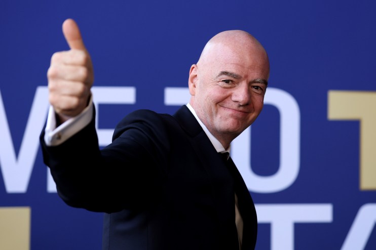 Gianni Infantino smiling and giving a thumbs up, wearing a suit against a blue and white background.