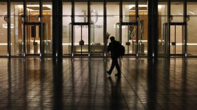 Silhouetted person walks past a modern building with 10 Upper Bank Street visible on its glass facade at night.