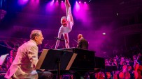 Carsten Windhorst performing with a gymnast, pianist, and orchestra under purple lights.