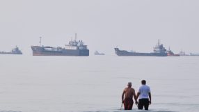 Two men stand in the ocean with multiple oil tankers and cargo ships in the hazy distance.