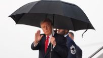 Donald Trump, in a suit and red tie, waves while holding a black umbrella, with a man in military uniform behind him.