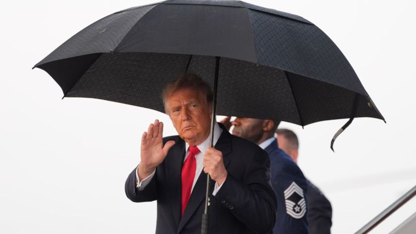 Donald Trump, in a suit and red tie, waves while holding a black umbrella, with a man in military uniform behind him.