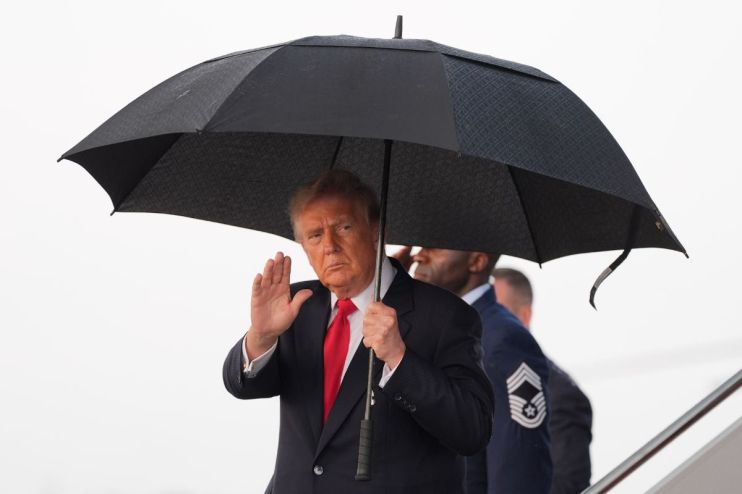 Donald Trump, in a suit and red tie, waves while holding a black umbrella, with a man in military uniform behind him.