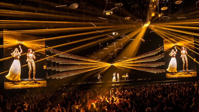ABBA Voyage concert with holograms of Agnetha and Frida performing, golden lights, and a live audience.