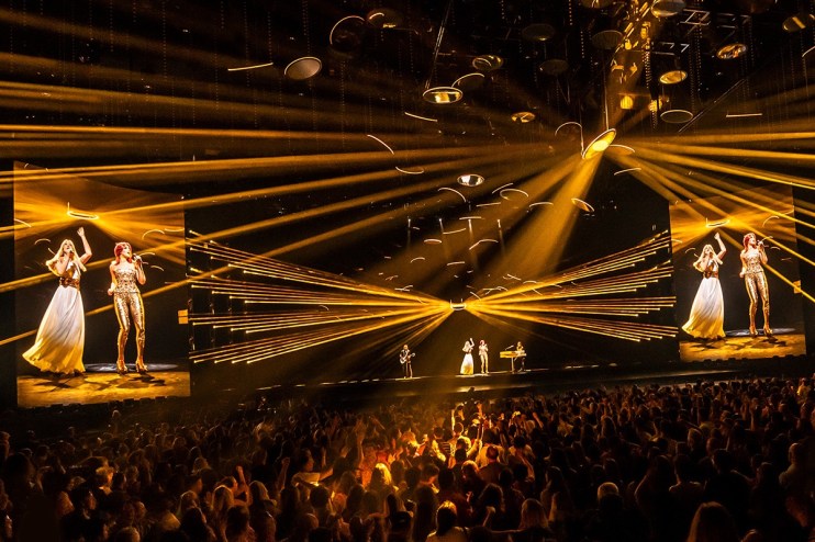 ABBA Voyage concert with holograms of Agnetha and Frida performing, golden lights, and a live audience.