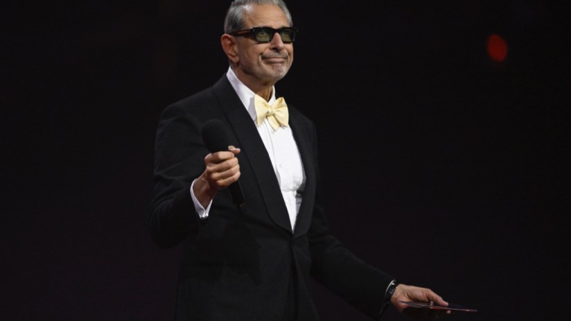 Jeff Goldblum in a black tuxedo with a yellow bow tie and dark sunglasses, holding a microphone, smiling.