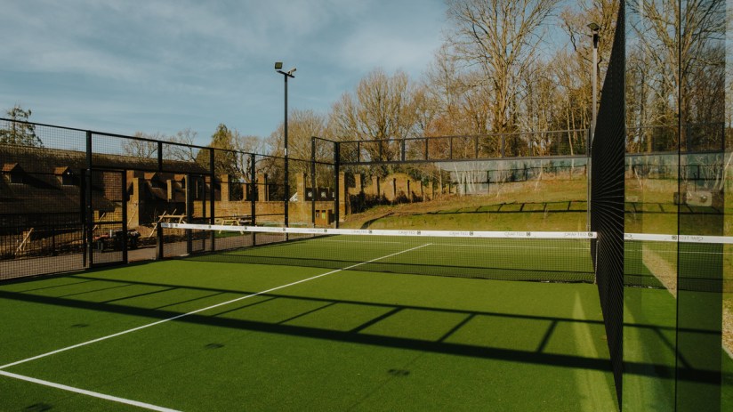 Outdoor padel tennis court with green turf, high fences, and a net, against a backdrop of trees and buildings.