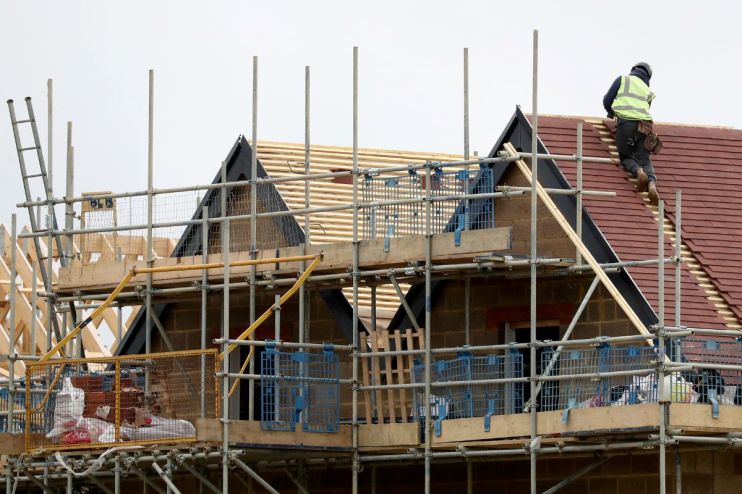 Construction worker in high-visibility vest on a new house roof with red tiles, surrounded by scaffolding.