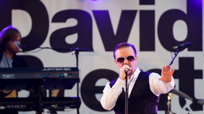 Ricky Gervais as David Brent performing on stage, wearing sunglasses and a patterned vest.
