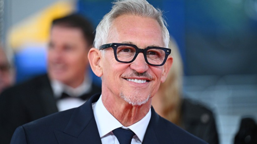 Gary Lineker smiling in a dark suit, white shirt, and navy tie, wearing black-framed glasses.