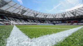 Low-angle view of a football stadium from the pitch, showing the corner marking and empty seats under a blue sky