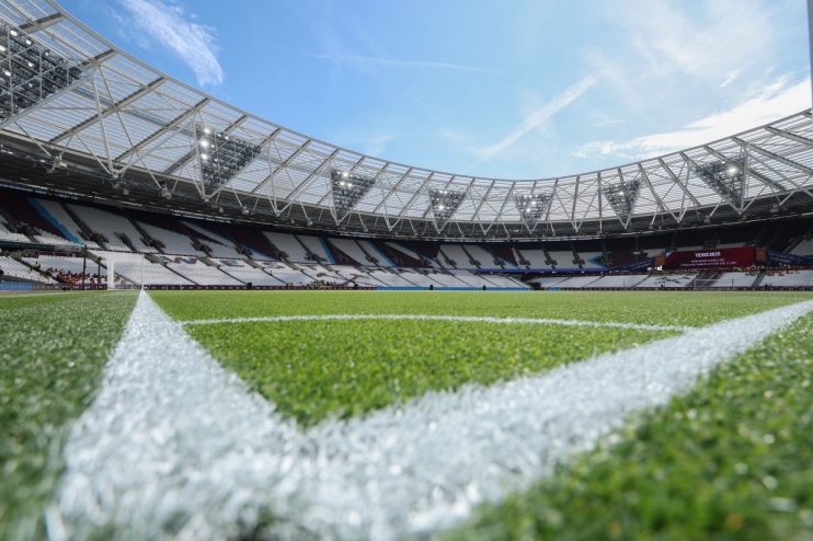 Low-angle view of a football stadium from the pitch, showing the corner marking and empty seats under a blue sky