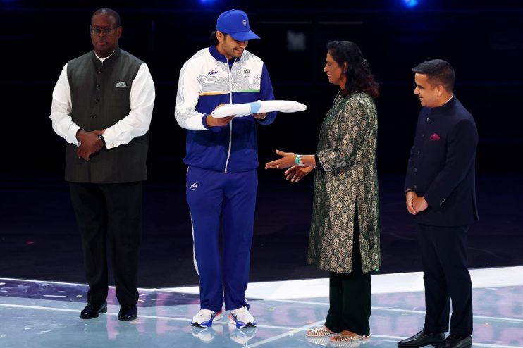 Indian athlete in blue tracksuit receives a white scroll from a woman in a green traditional dress, flanked by two men in ...