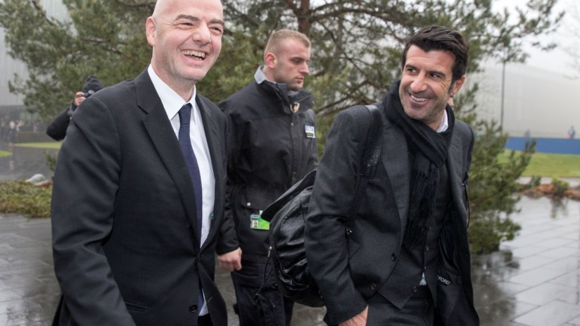 Gianni Infantino and Lu&iacute;s Figo smiling, walking outdoors with security, on a wet day.
