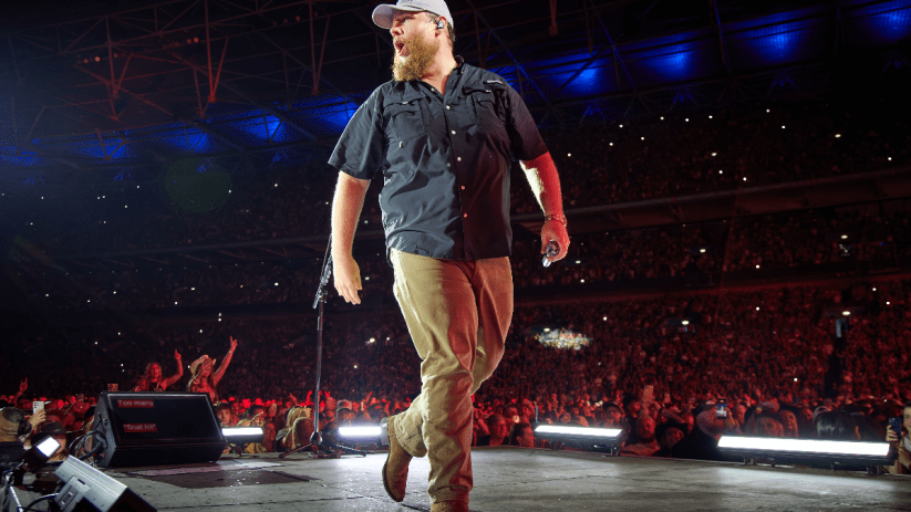 Luke Combs performing on stage at Wembley Stadium, facing right with a microphone, large crowd visible