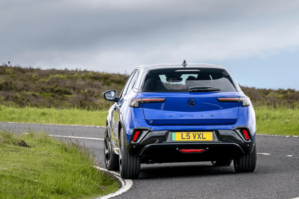 Rear view of a blue Vauxhall Mokka driving on a winding road with green hillsides under a cloudy sky.