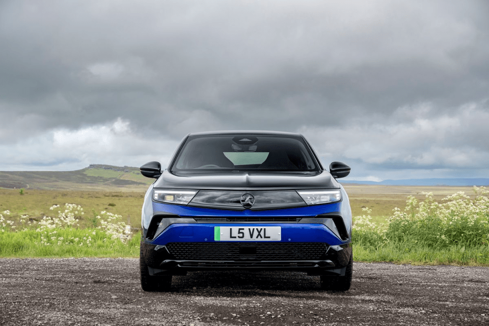 Front view of a blue and black Vauxhall Mokka parked on a gravel road with a cloudy sky and green hills in the background.