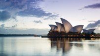 Sydney Opera House at dawn with illuminated sails reflecting on calm water and a cloudy sky