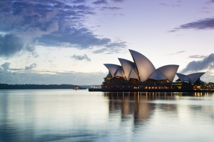 Sydney Opera House at dawn with illuminated sails reflecting on calm water and a cloudy sky