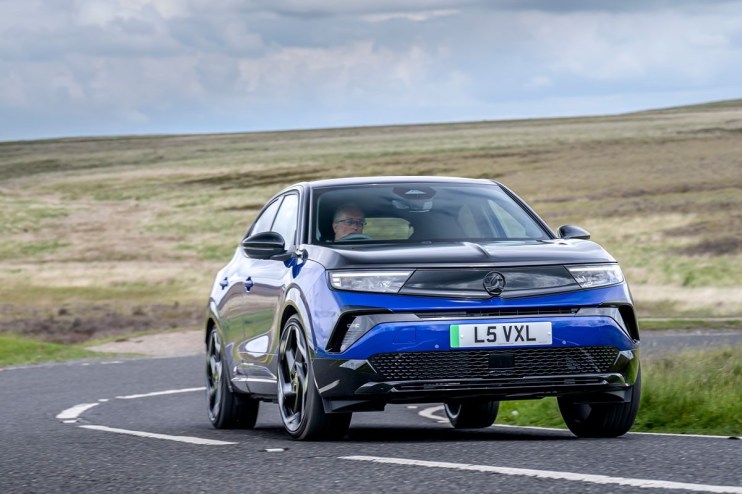 Blue and black Vauxhall Mokka driving on a winding country road with a driver visible, against a hilly landscape.